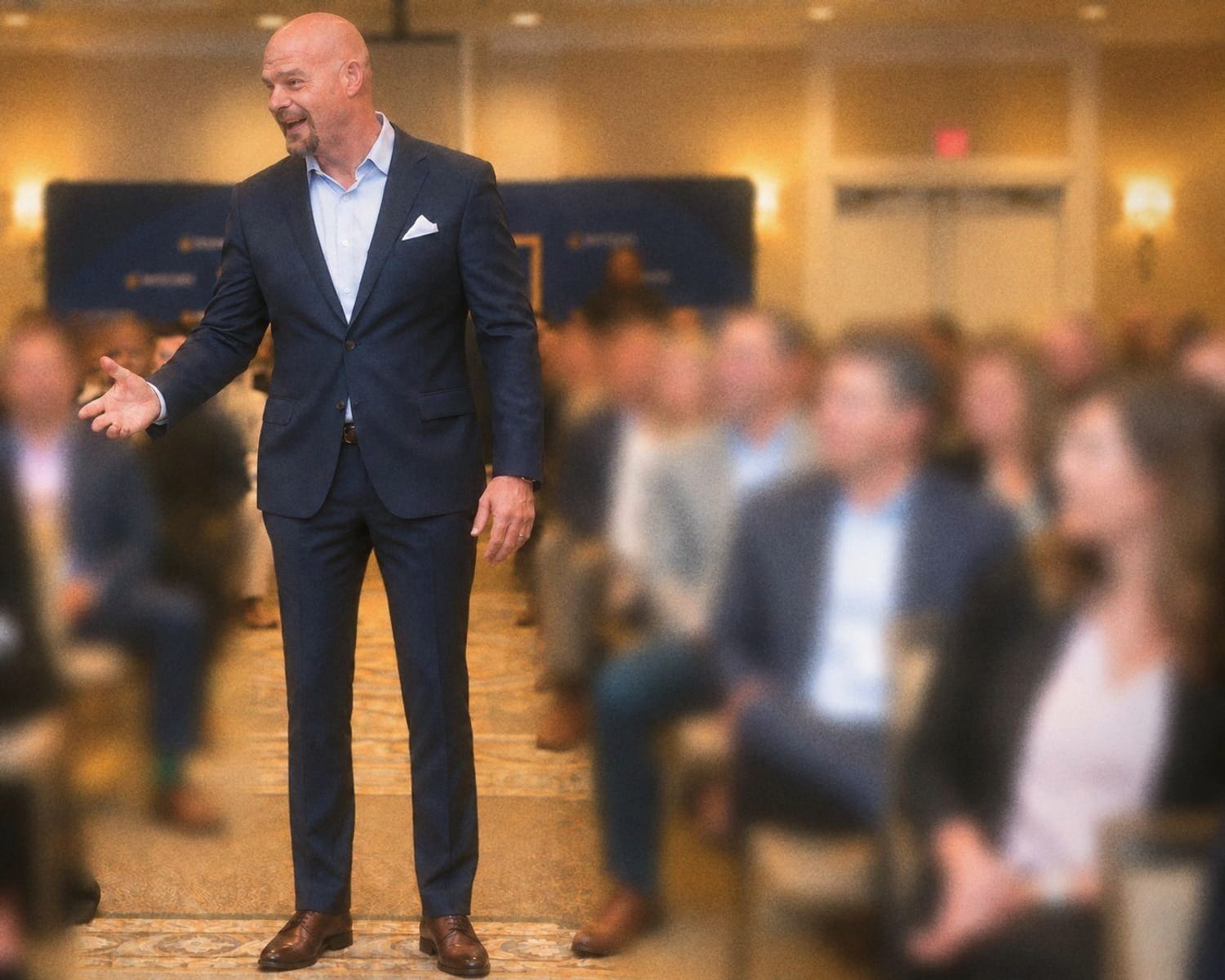 Dean Eric Smith in a navy suit, laughing and reaching toward executives seated along the aisle