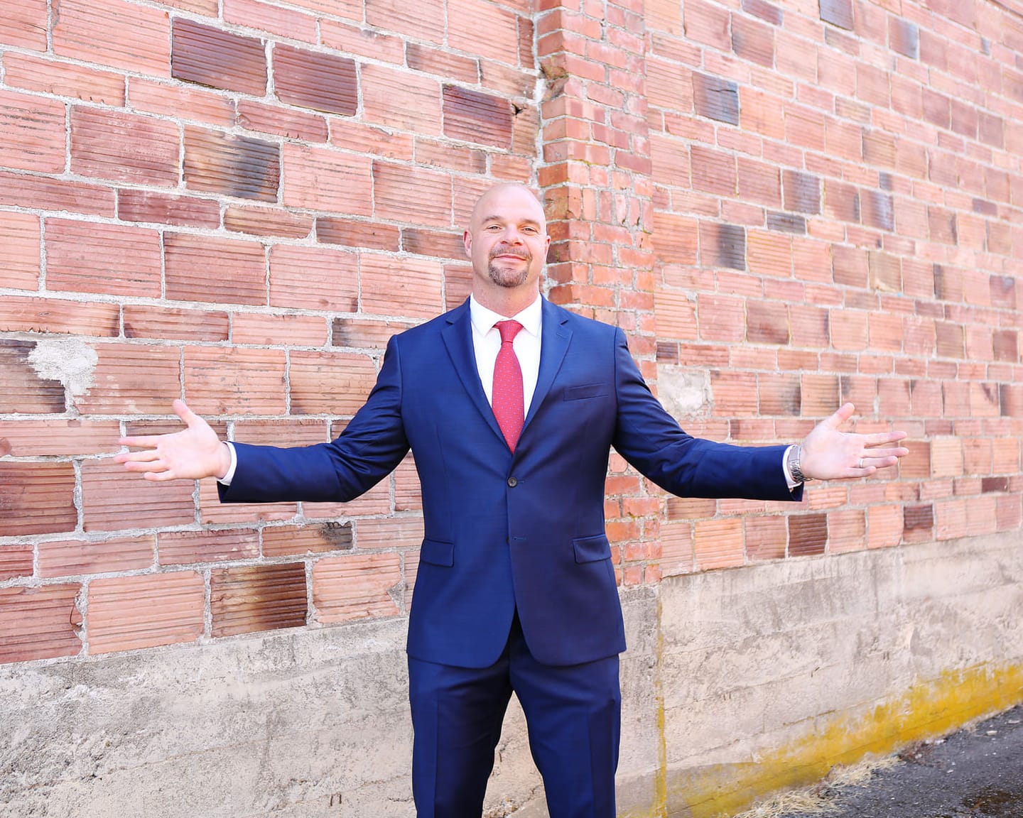 Dean Eric Smith in a navy suit and red tie, arms open wide, in front of a red brick wall