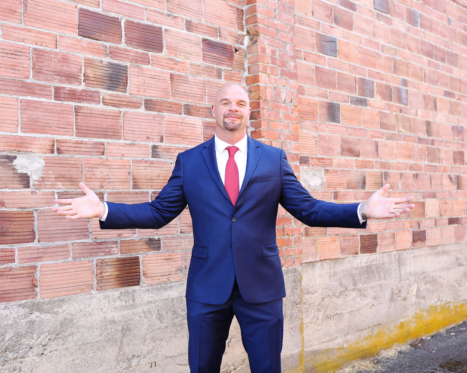 Dean Eric Smith in a navy suit and red tie, arms open wide, in front of a red brick wall